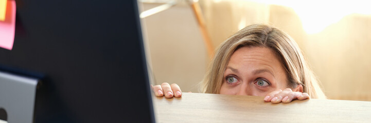 Woman hides behind office desk and looks at computer monitor.