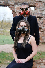 Two people in black clothes and gas masks pose against the background of a destroyed brick wall. Steampunk goggles on a man