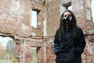 Post apocalypse female survivor. Young pandemic survivalist woman in gas mask looking in front of her on the background with apocalyptic war area around. Sad woman in breathing oxy mask.