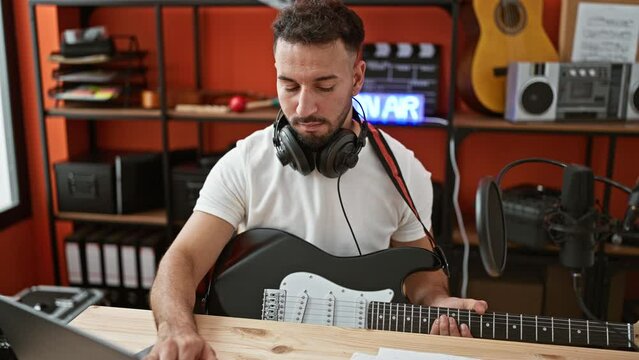 Young arab man musician playing electrical guitar composing song using laptop at music studio