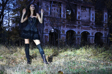 A mysterious woman in a short black dress with a corset, Halloween makeup and a black veil poses at night against the backdrop of an old abandoned building