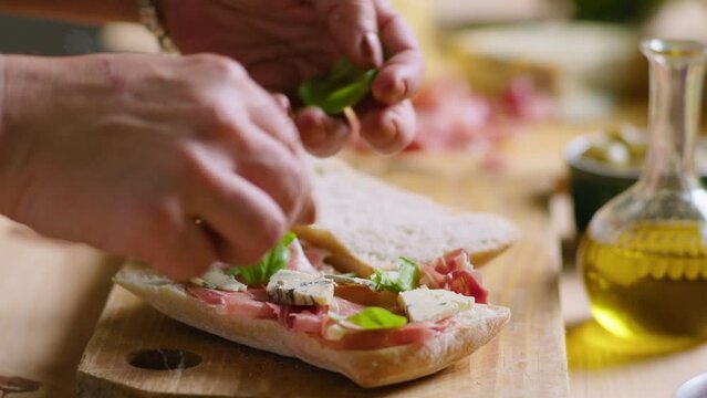 Hands of woman adding fresh basil leaves to gourmet sandwich with prosciutto and blue cheese on ciabatta bread. Close-up view, selective focus