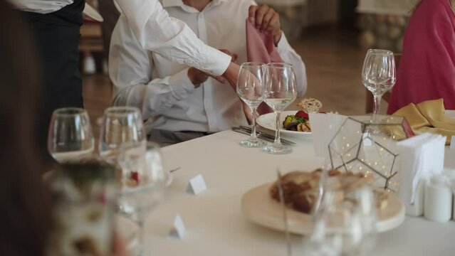 At festive table man puts napkin on his feet and waiter puts dish on table. Serving in restaurant, blurred woman unlocked smartphone, takes pictures of food. There are empty glasses on served table.