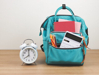 Front view  of green backpack with school supplie and white vintage alarm clock 8 o'clock on wooden table and white  background with copy space.
