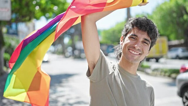 Young hispanic man smiling confident holding rainbow flag at street
