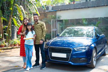 Indian family standing with new car. girl holding puja thali in hand.