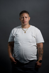 A woman in a white t-shirt poses for a casual portrait. She is in a studio, in front of a gray backdrop. Adding backstory, she is lesbian, with a butch aesthetic. She has a fade haircut and is Black.