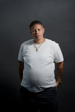 A woman in a white t-shirt and gold necklace poses in a gray studio. Adding more backstory, she is African American, and has a self-identified butch lesbian aesthetic.