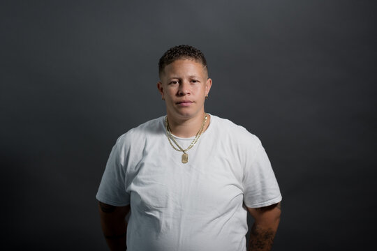 Wearing a white tee and a gold necklace, a woman poses for a portrait against a gray backdrop. She is confident and assertive. With pride in her lesbian identity, her style has a distinct butch edge.