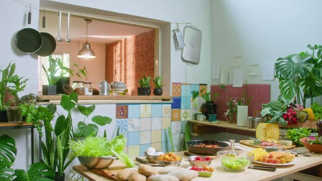 Interior of domestic kitchen decorated with lots of green plants, fresh vegetables and assorted food ingredients on the table. Zoom out shot, no people