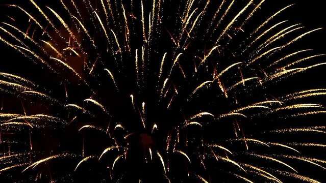 Slow-motion shooting of large balls of festive fireworks disintegrating into flickering golden rays and smoke in the black night sky.