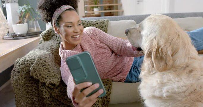 Happy biracial woman taking photo with golden retriever dog using smartphone at home, slow motion