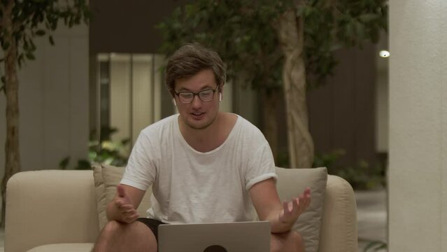 Emotional man sitting at home on sofa, waving hand and gesturing while talking on a video call, virtual meeting with friends and family. Concept of distance, communication and internet