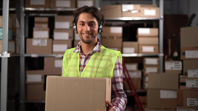 Male warehouse worker wearing headset with a large box looking at camera and smiling