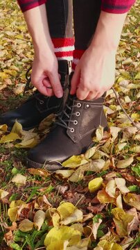 Close-up of an unrecognizable woman tying shoelaces on black leatherette boots in yellow leaves in autumn outdoors, vertical video