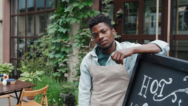 Slowmo medium portrait of male African American cafe worker standing next to blackboard with special offer written on it outdoor at daytime