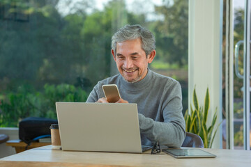 Fototapeta premium mature asian man working remotely on internet online over laptop and smartphone technology vacation,take a break in cafe at resort garden,destination in Thailand.concept of travel freely,digital nomad