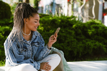 African american woman using mobile phone while sitting in park - obrazy, fototapety, plakaty