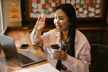 Asian woman smiling and waving hand while making video chat via laptop in cafe
