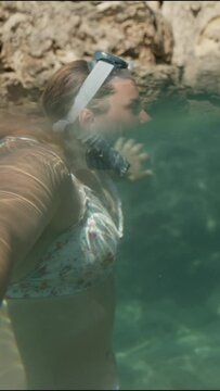 Vertical video. The camera captures dual perspectives above the water, a young woman wearing a snorkeling mask and breathing tube; below the water, underwater cliffs can be seen.