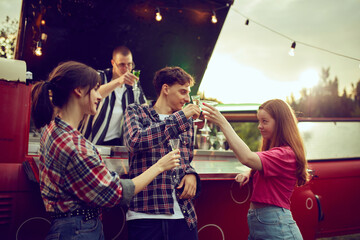 Young cheerful people, friends standing in van with guy making delicious cocktails in it. Good time at outdoor party. Concept of friendship, leisure time, weekends, summer, party