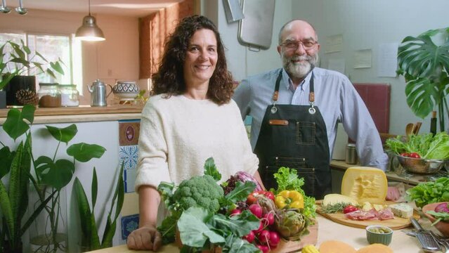 Happy senior man in apron and his wife standing together by kitchen table with fresh vegetables and cheese, posing together for the camera and smiling. Video portrait, medium shot