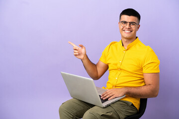 Young man sitting on a chair with laptop pointing finger to the side