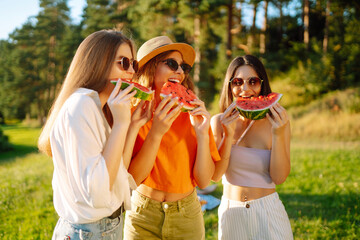 Fototapeta premium Group of smiling best friends have fun and eat watermelon on a hot summer day at a picnic. Three friends are relaxing and enjoying nature. The concept of vacation, picnic, friendship.
