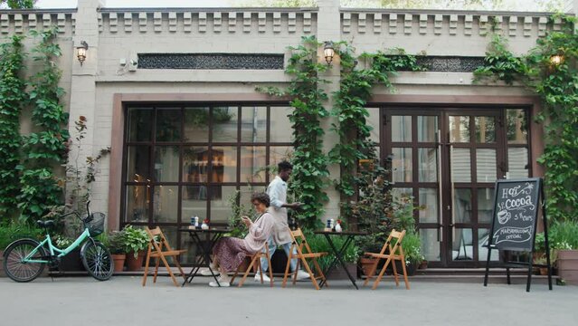 Establishing shot of Caucasian woman sitting at table in outdoor cafe and African American waiter coming inside at daytime