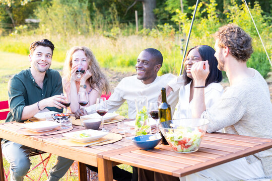 A vibrant and diverse evening garden party, where a multi-ethnic group of millennial friends gathers around the dinner table, engaging in lively conversations and enjoying each other's company