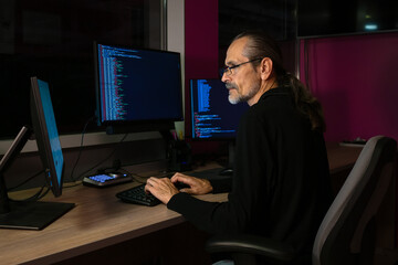 A middle-aged programmer with glasses and a ponytail works with code at a workplace with three monitors in a darkened office