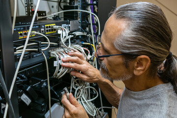 A middle-aged engineer with glasses and a ponytail plugs in network cables at the back of a rack in a server room