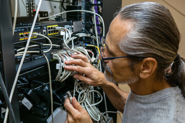 A middle-aged engineer with glasses and a ponytail plugs in network cables at the back of a rack in...