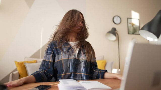 Happy young smiling woman sings and dances sits at desk turning head shaking hairs. Carefree girl student at working place with laptop enjoys her life having break in classes in distance studying.