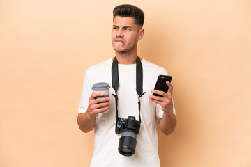 Young photographer caucasian man isolated on beige background holding coffee to take away and a mobile while thinking something