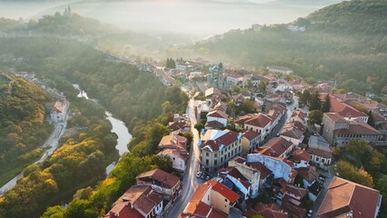 Aerial shot of sunny slightly foggy morning in Veliko Tarnovo, Bulgaria. Flying over old houses, Ascension Cathedral and river in the canyon in Veliko Tarnovo 2 - 628453671