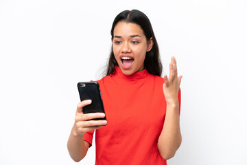 Young Colombian woman isolated on white background looking at the camera while using the mobile with surprised expression