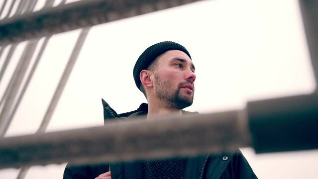 Sad depressive man standing on bridge waiting somebody with backpack looking ahead in winter snowy weather. Bearded young guy in warm clothes in city urban area. Meeting on street in cold weather.