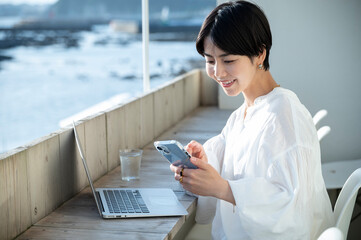 Asian woman using a smartphone and laptop on the terrace of a cafe on the coast