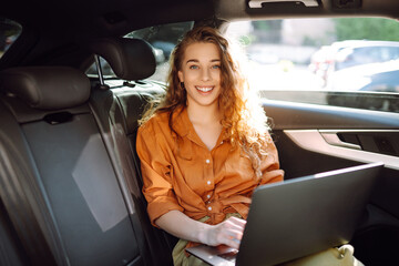 Business beautiful woman with a modern laptop sits in the back seat of a car. Successful startup, remote work. Freelance. 
