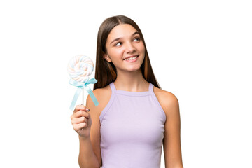 Teenager caucasian girl holding a lollipop over isolated background looking up while smiling