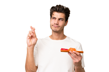 Young caucasian man holding sashimi over isolated background with fingers crossing and wishing the best