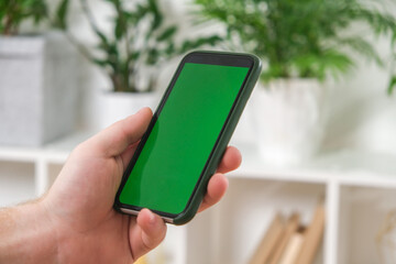A man holds a phone with a green screen in his hands against a background of green house plants. Mockup. Chromakey.