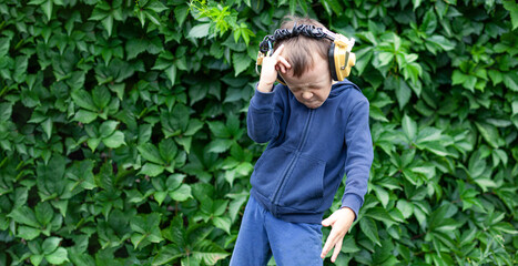 A positive little boy of 6 years old in yellow retro headphones listens to music and dances. A boy dances against a wall of plants. Banner with space for text