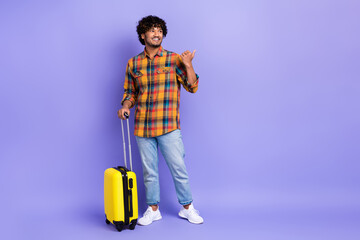 Full body photo of young guy wear trendy jeans direct finger mockup schedule airport carry suitcase isolated on violet color background