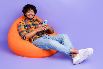 Full length photo of optimistic man dressed shirt sit in orange pouf hold smartphone read post...