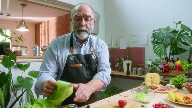 Senior food blogger with gray hair and beard taking tomato and cutting lettuce, preparing vegetable salad and talking on camera, giving online culinary class or filming vlog in the kitchen
