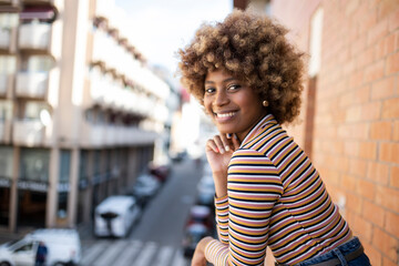 Portrait of smiling woman standing in a balcony with city background. Girl staring at camera with a smile in a high window.