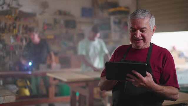 Happy senior worker supervising carpentry business while using tablet device to receive online orders. Portrait of Small business owner of local shop wearing apron
