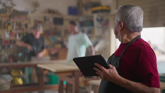 Happy Business owner of carpentry Local Store workshop in Apron Using Tablet supervising workers in background with tools